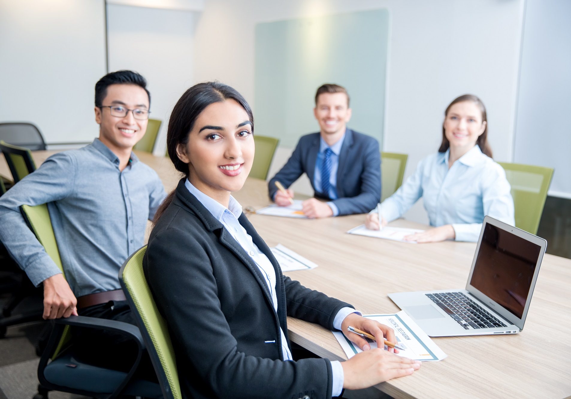 Four smiling middle-aged multi-ethnic business people turning to camera, working and discussing ideas while sitting at big table in conference room. Side view.